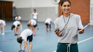 Portrait of happy female physical education teacher at school gym. Young happy coach using stopwatch during PE class at school gym and looking at camera. Her students are exercising in the background.